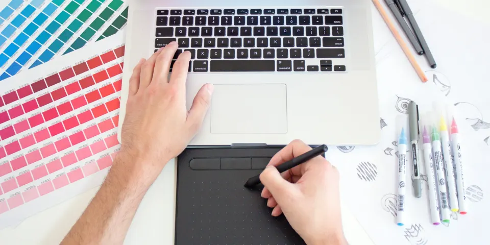 Manos de un diseñador trabajando en un portátil y una tableta gráfica junto a muestras de color