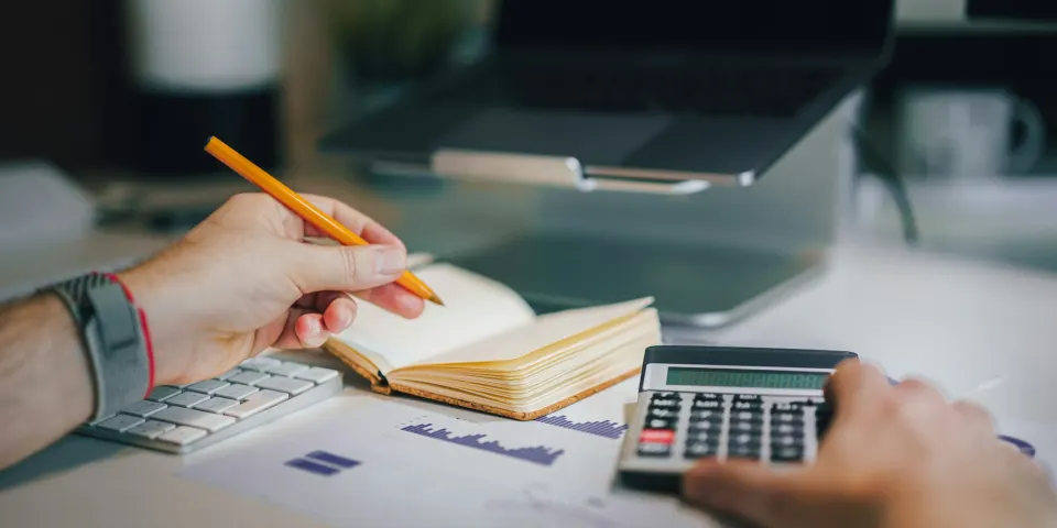 Persona haciendo cuentas con calculadora y cuaderno junto a un computador portátil