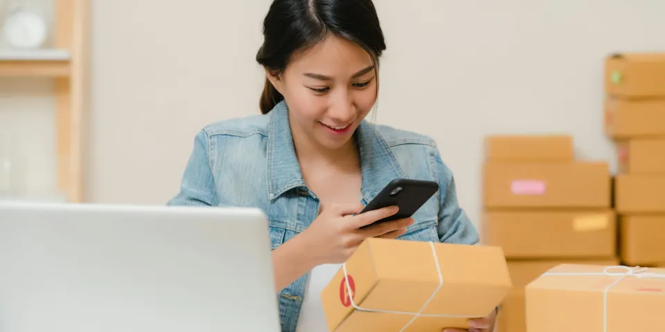 Emprendedora revisando pedidos en su celular rodeada de cajas listas para enviar